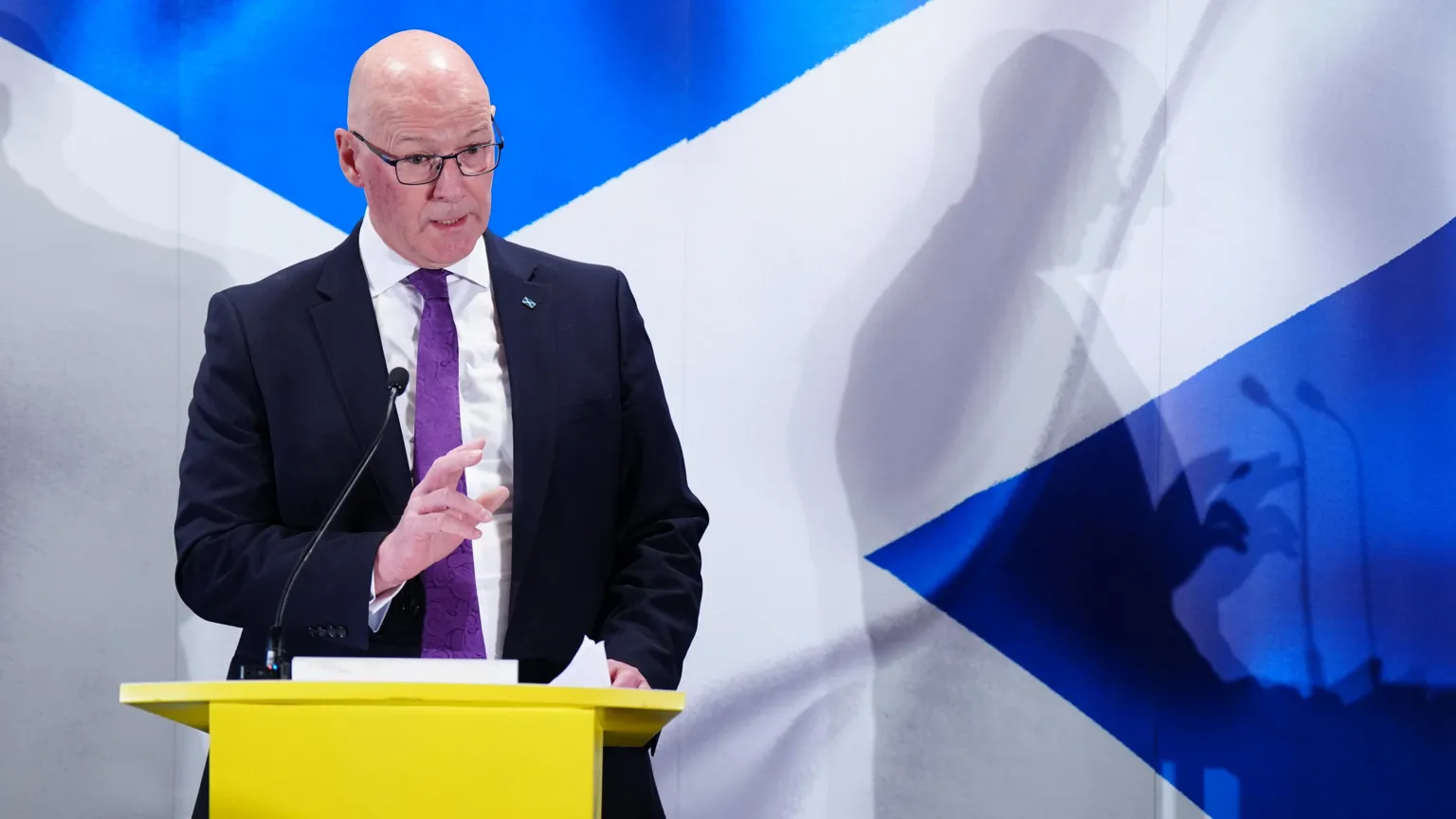 John Swinney standing at a yellow lectern with a saltire behind him.