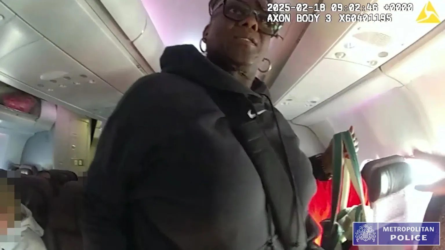 Metropolitan Police A low-angle body-cam shot inside an airplane aisle, showing a Janice Nix in a black hooded sweatshirt and glasses holding a striped tote bag. A Metropolitan Police logo is in the corner.