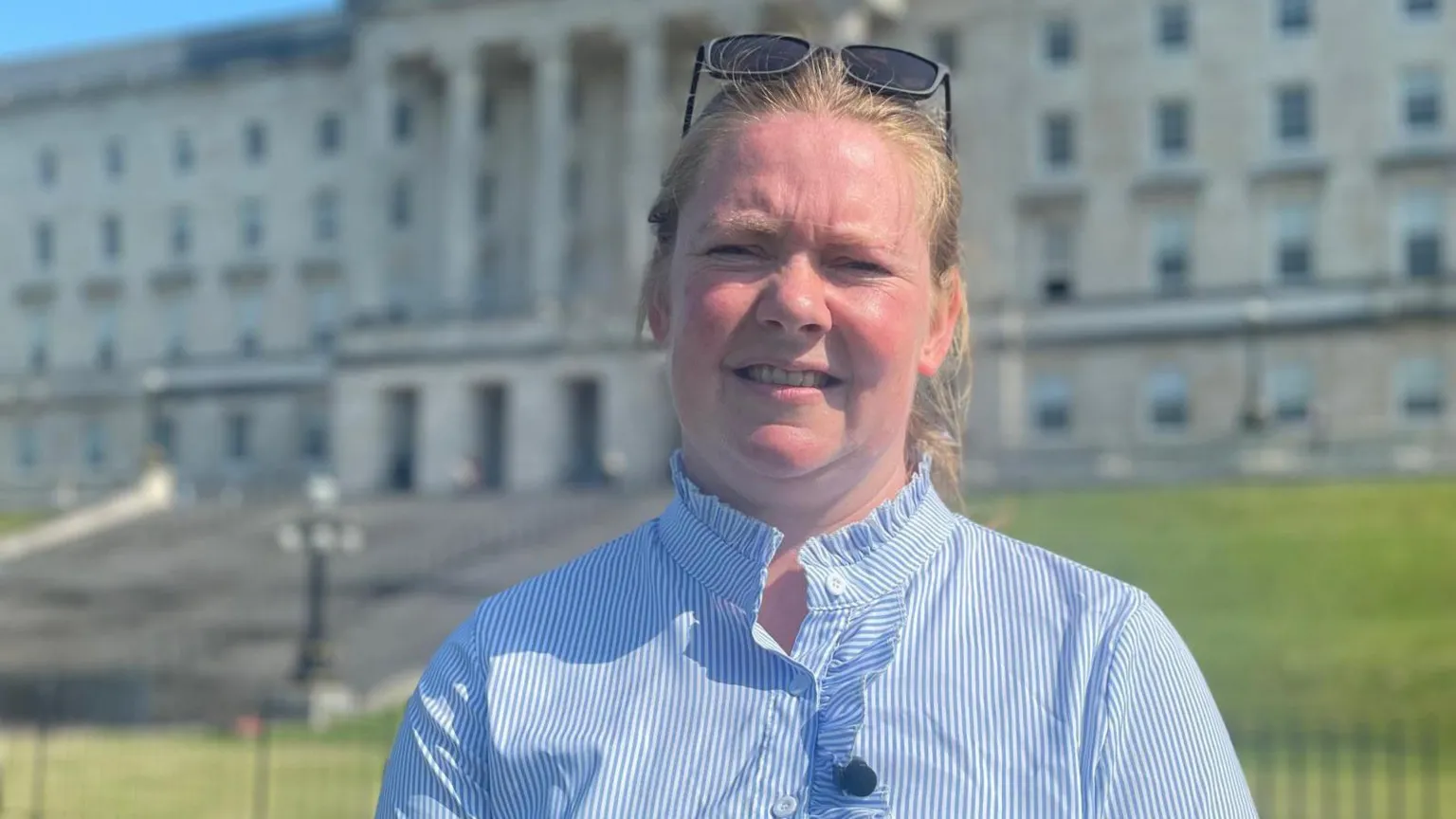 A woman, with blonde hair tied back, wearing a blue and white striped shirt and sunglasses on her head. She is standing in front of the Stormont building. 