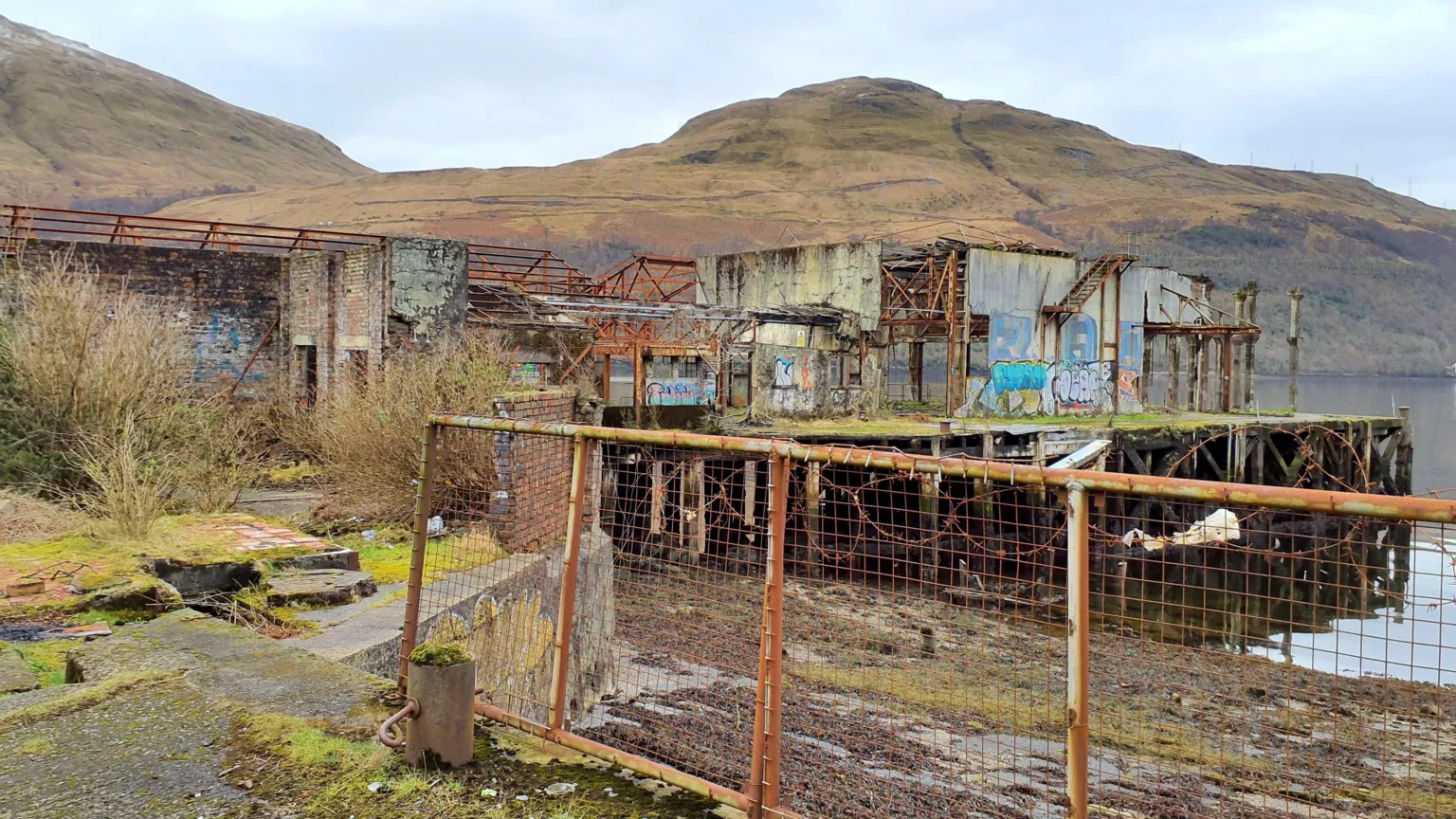 Loch Lomond & The Trossachs National Park Authority A view from outside the building of the former torpedo station. It is rusting and covered in graffiti. The building sits on a loch, with hills in the distance.