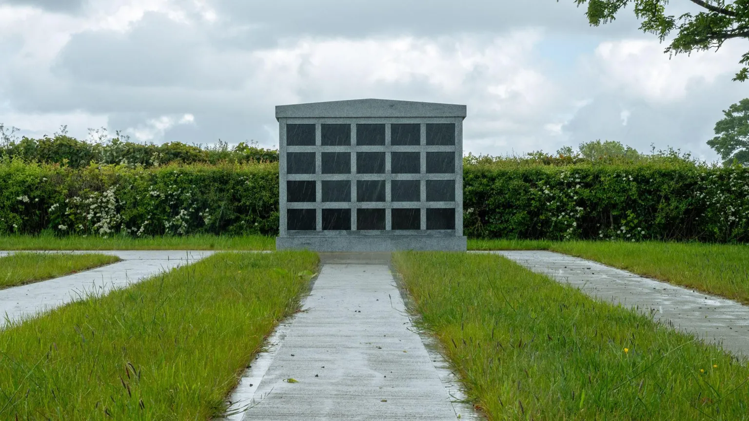 DCSDC A grey columbarium with a number of rows in a housed space. It is near a grassy area with footpaths and shrubbery.