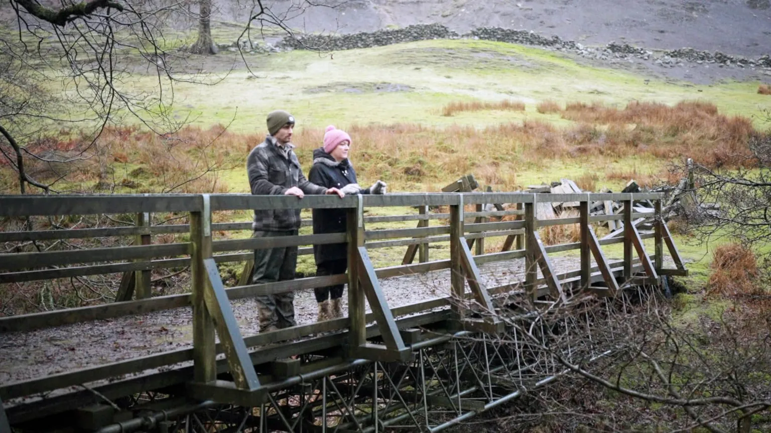 Nathan’s mum Jess and her partner Andrew stand together on a small wooden footbridge, named after Nathan, over a narrow stream in a quiet rural landscape. They’re bundled in warm coats and hats and around them is open countryside with grass, bare trees, and a low stone wall in the distance.