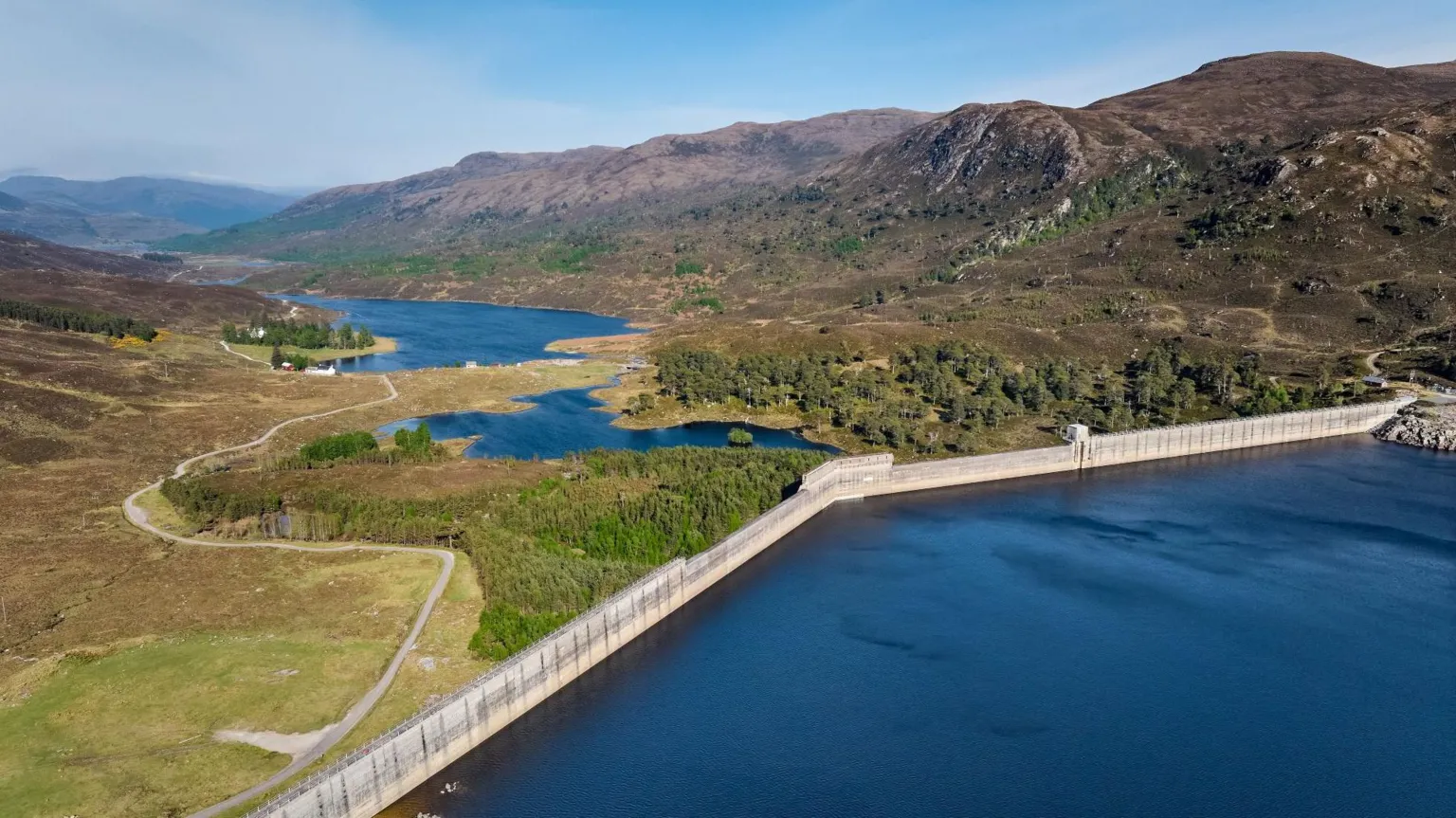  A dam holding back the waters of Loch Mullardoch in the foreground with trees and a receiver loch in the distance.