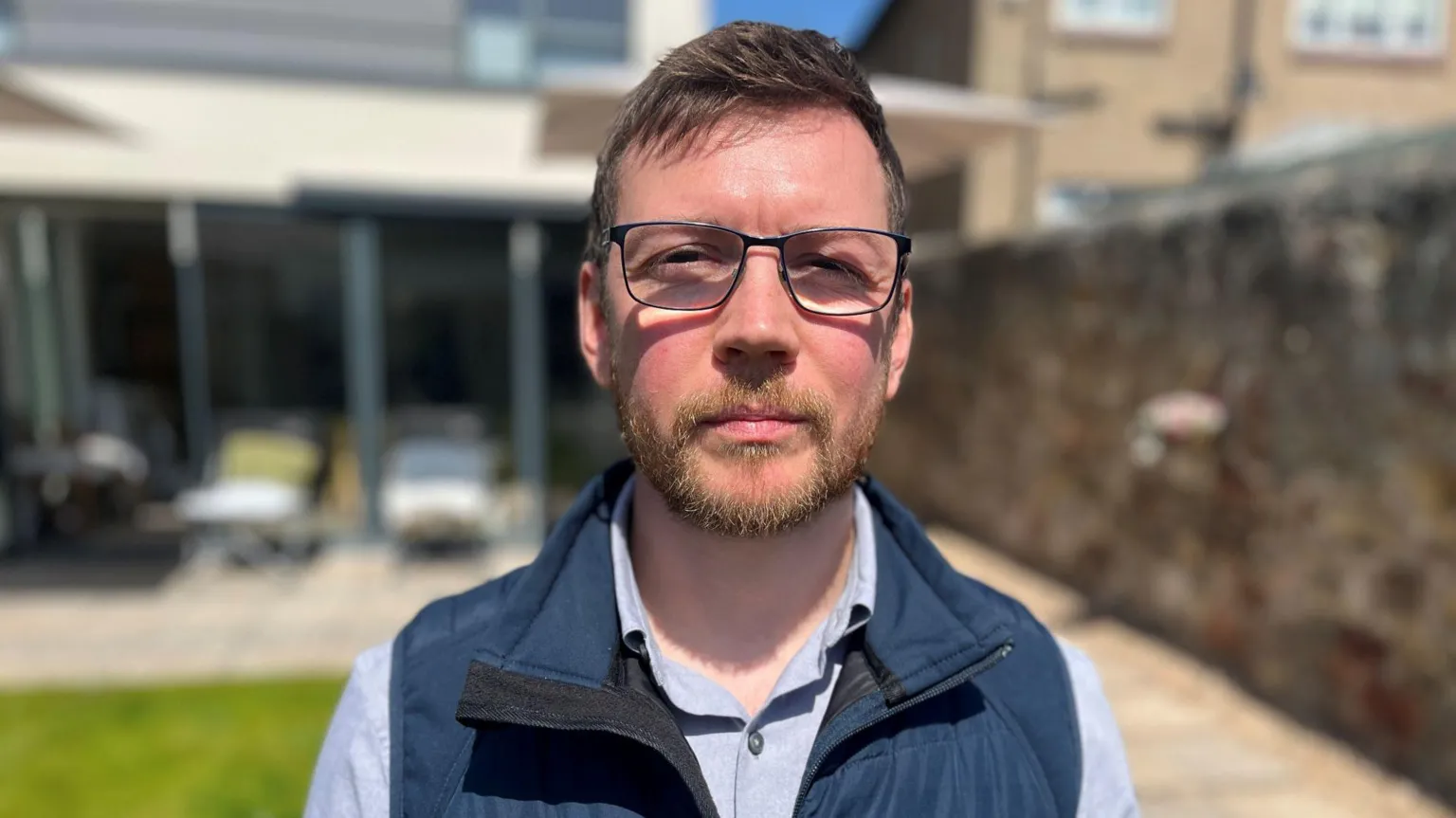 Josh King, in a blue body warmer, with a brown beard and hair, wearing black glasses. He is standing in a residential garden.