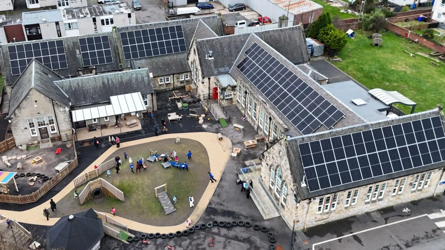 An aerial image of a school with multiple buildings and a central play area. There are children in the playground and several of the roofs are covered with solar panels.