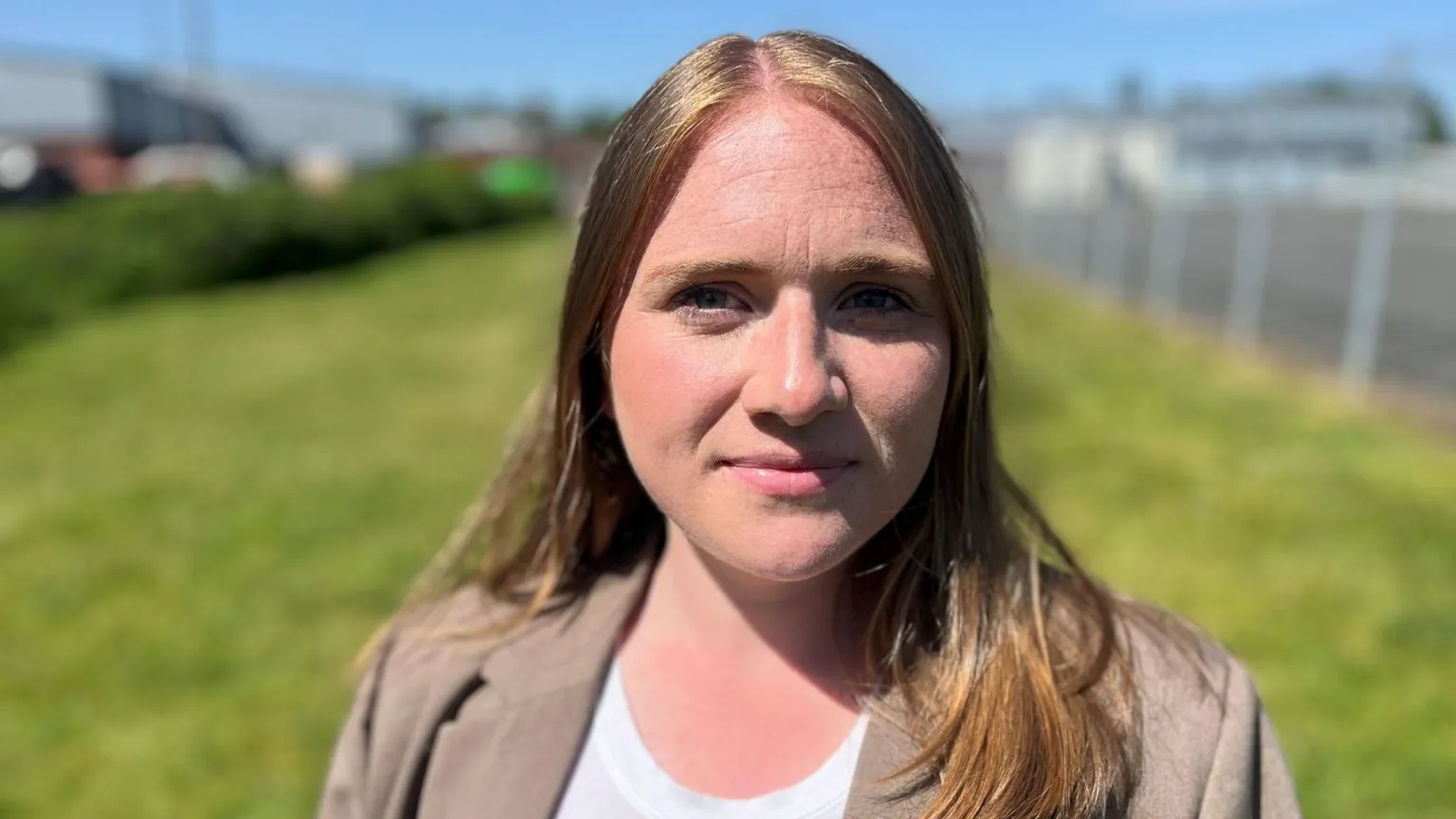 Laura young with long auburn hair. She is standing on grass and wearing a brown jacket over a white top.