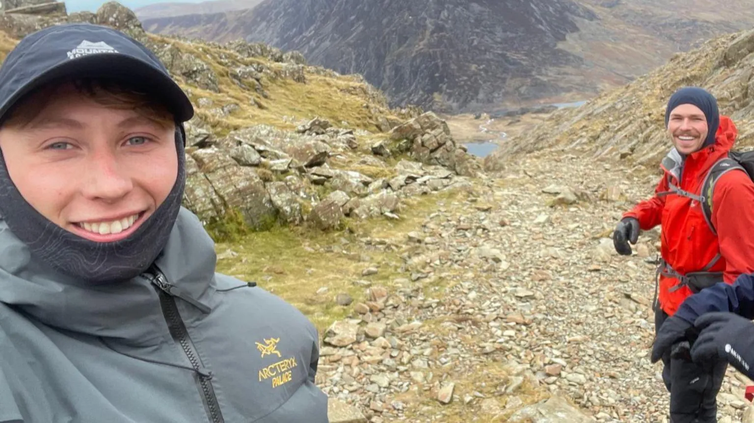 Matty Belcher | Brandan Smith A man with a dark blue baseball cap and grey windbreaker jacket smiles at the camera. To his left stands a man in a red jacket and blue hood, with a large hiking backpack. Behind them is a rocky grey mountain and blue lake in the valley below.