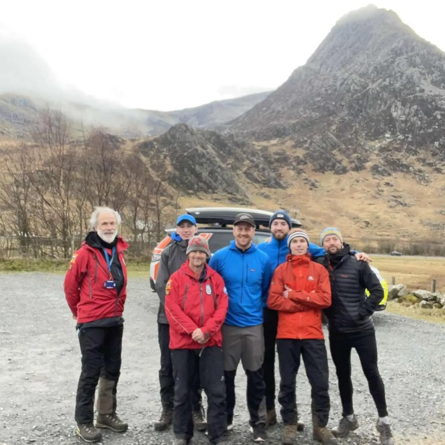 Matty Belcher | Brandan Smith A group of men in red, blue and orange winter hiking jackets stand with their arms around each other beneath a rocky grey mountain.