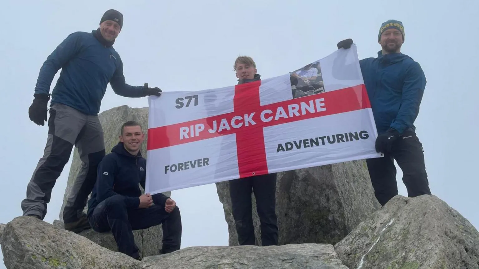 Matty Belcher | Brandan Smith Four men stand on top of large grey rocks holding up a st George's cross flag which reads 'RIP Jack Carne'