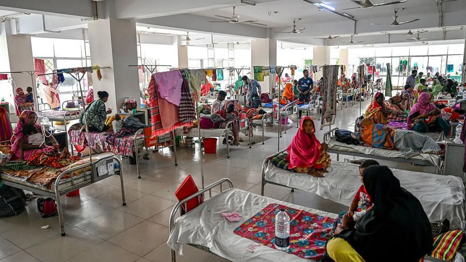  A crowded hospital ward with women sitting cross-legged on their beds with colourful blankets. Some of the women have children