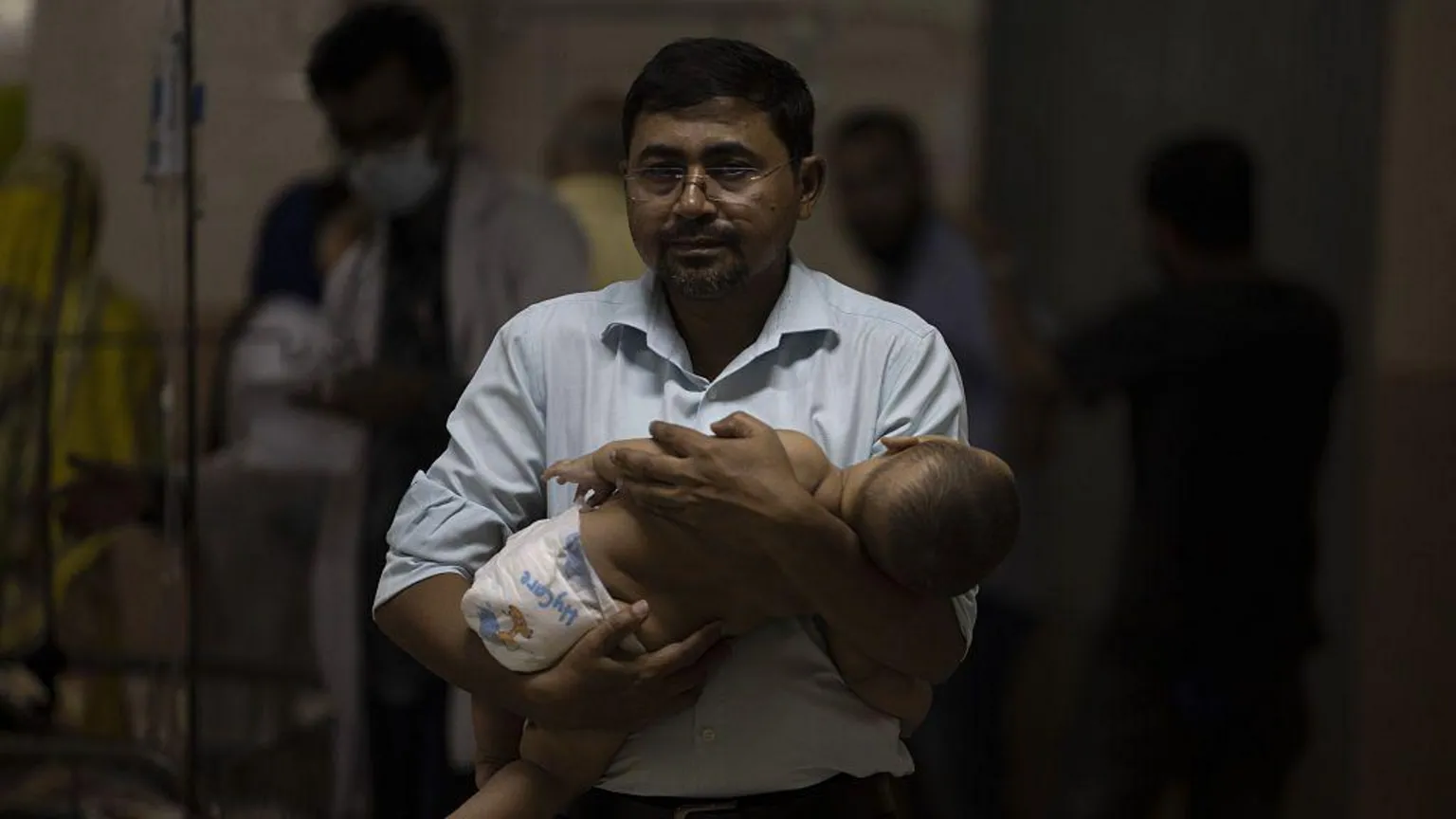  A man wearing a button-up shirt and glasses stands in a hospital holding a baby in his arms