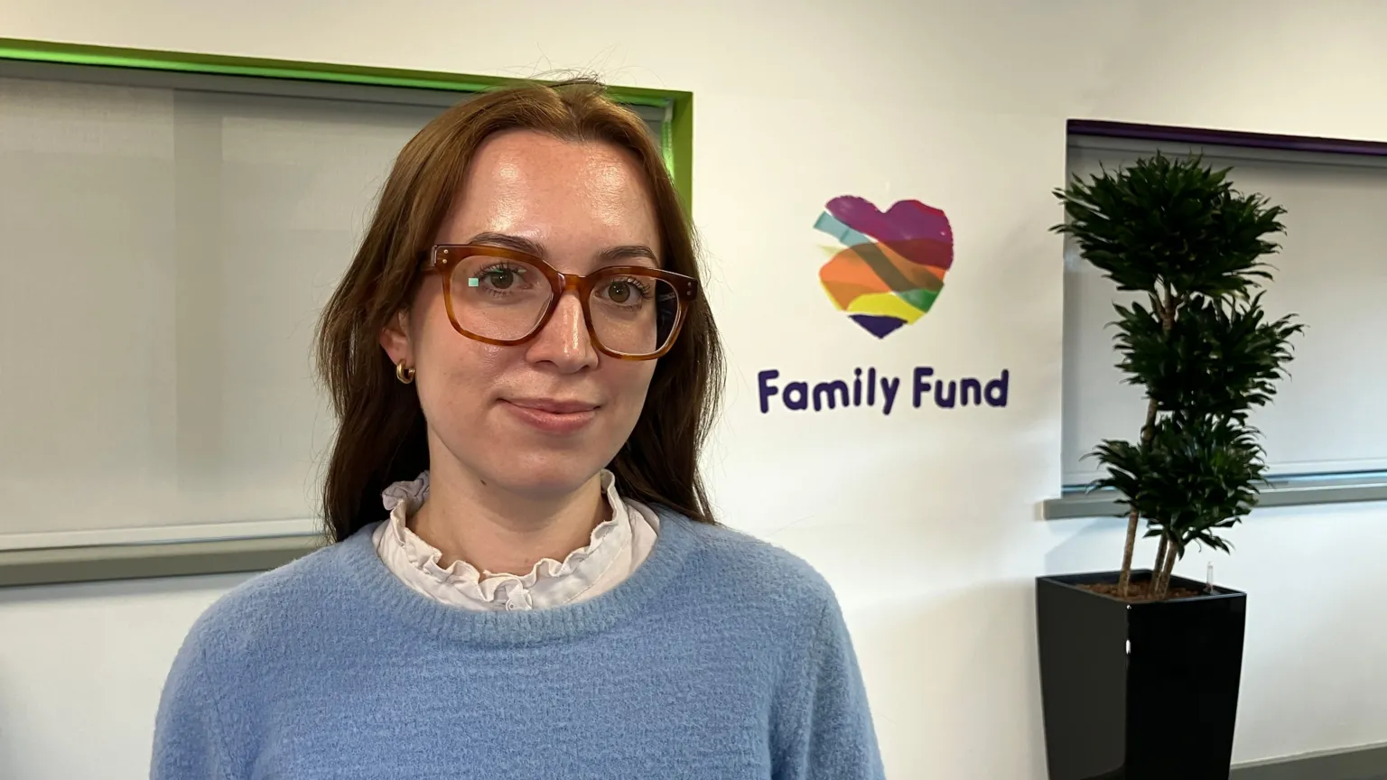 Kit Taylor/BBC A young woman with brown hair and glasses, a light blue jumper smiling at the camera in an office environment. In the background is a Family Fund logo and a potted plant. 