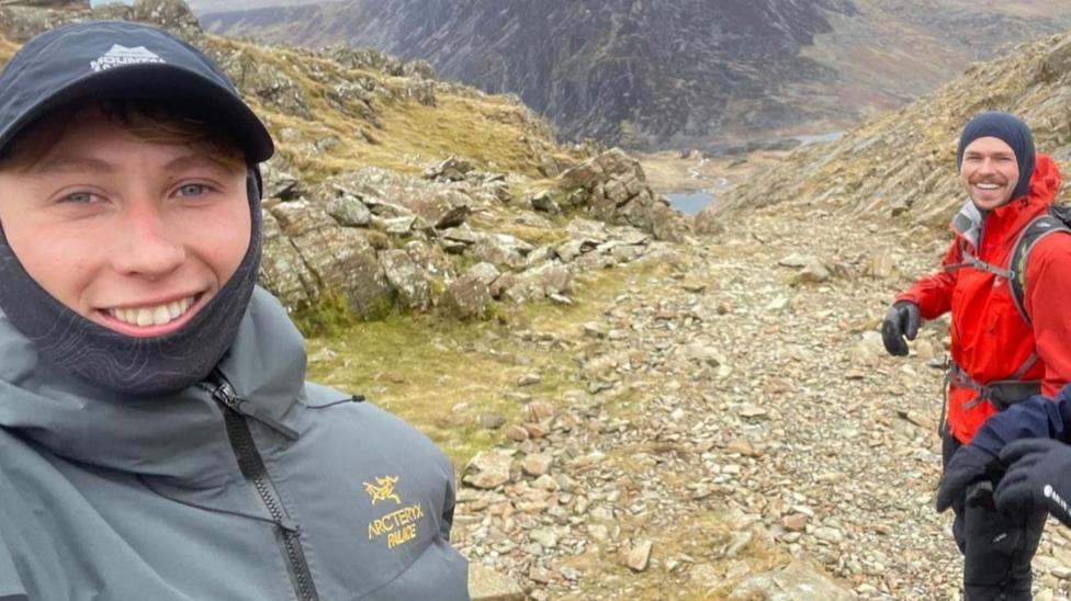 Mae dyn gyda het glas tywyll a siaced lwyd yn gwenu ar y camera. I'w chwith mae dyn mewn siaced goch, gyda sach gefn. Y tu ôl iddyn nhw mae mynydd llwyd creigiog a llyn glas yn y dyffryn islaw.