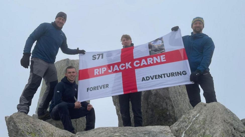 Pedwar dyn yn sefyll ar ben creigiau llwyd mawr yn dal baner sy'n nodi 'RIP Jack Carne', a 'forever adventuring'