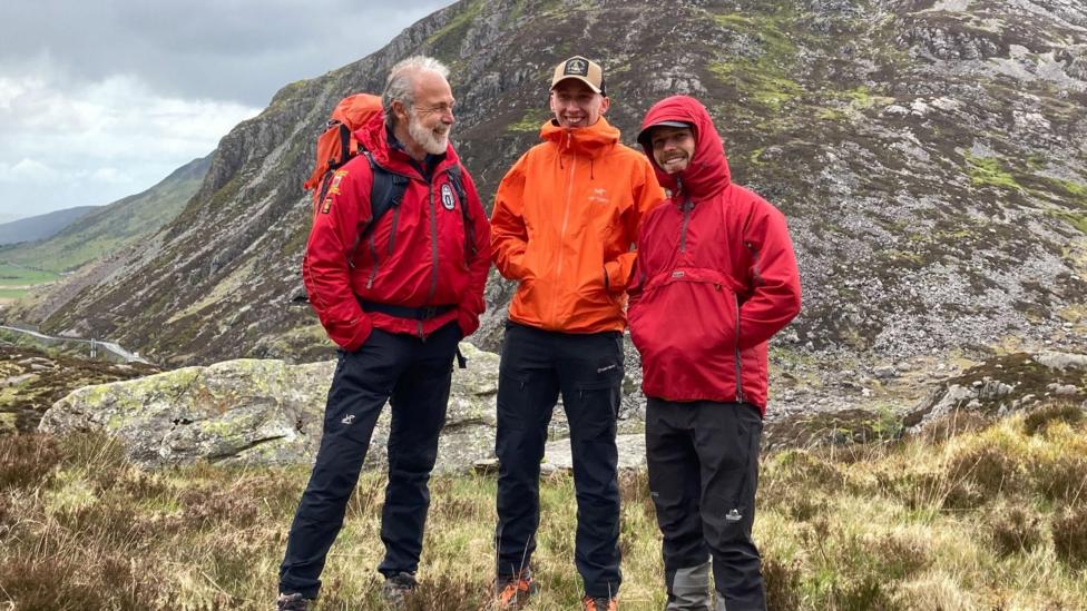 Mae tri dyn mewn siacedi coch ac oren yn sefyll yn gwenu ar ei gilydd o dan ochr mynydd