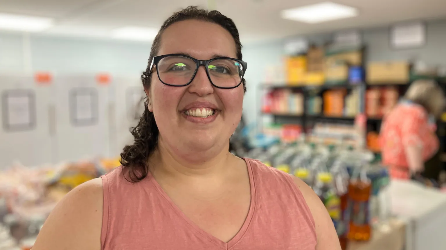 A woman with curly dark hair and glasses, wearing a pink v-neck vest top, is smiling at the camera. She is standing in a shop and their are bottled items behind her.