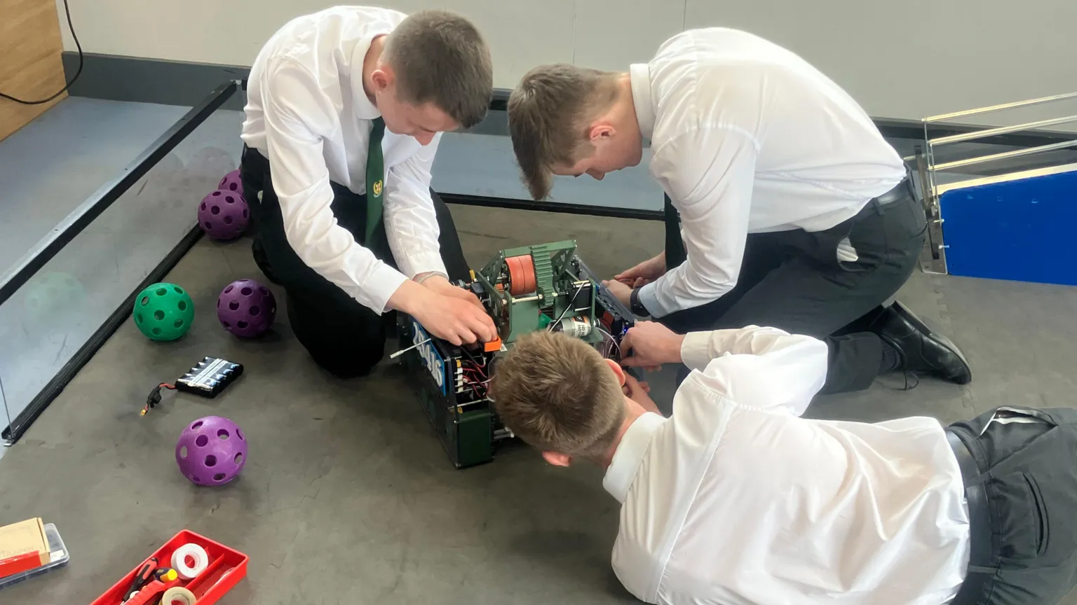 3 students wearing white shirts and black trousers sitting around the robot working on it.