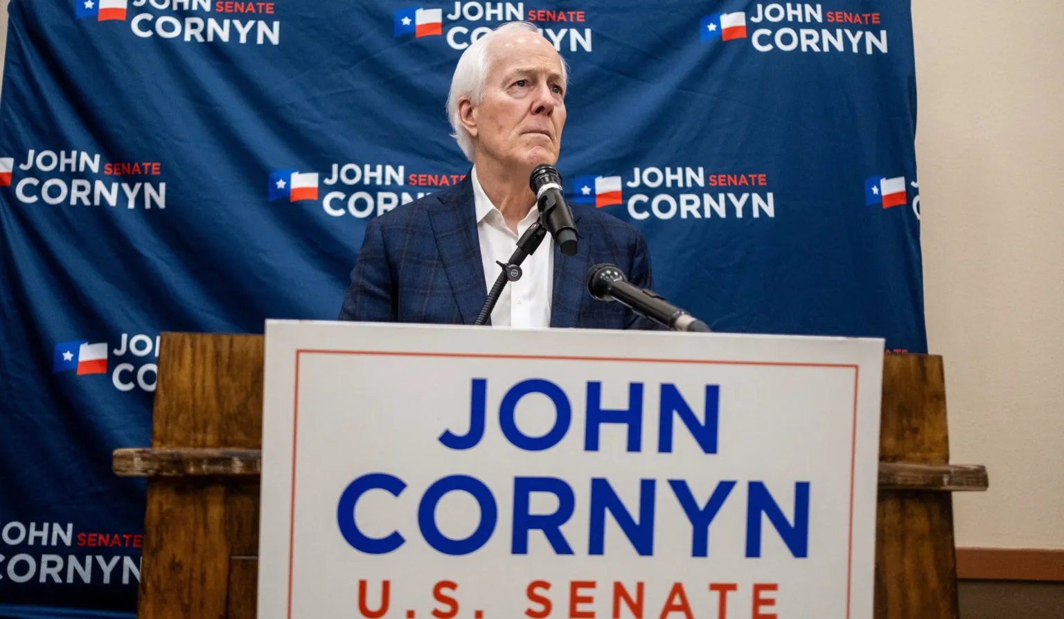 /Joel Angel Juarez John Cornyn stands at a podium with red white and blue campaign signs. His expression is serious and he is looking into the distance.
