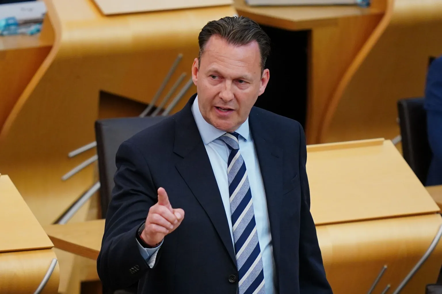 Scottish Conservative leader Russell Findally wearing formal business attire stands inside a modern parliamentary or legislative chamber with light wood desks arranged in rows. The individual is wearing a dark suit jacket, a light blue shirt, and a striped tie. One hand is raised with the index finger extended in a pointing gesture, suggesting emphasis during speech or debate. The background shows empty desks and chairs, softly lit with warm tones, indicating an indoor government or assembly setting.