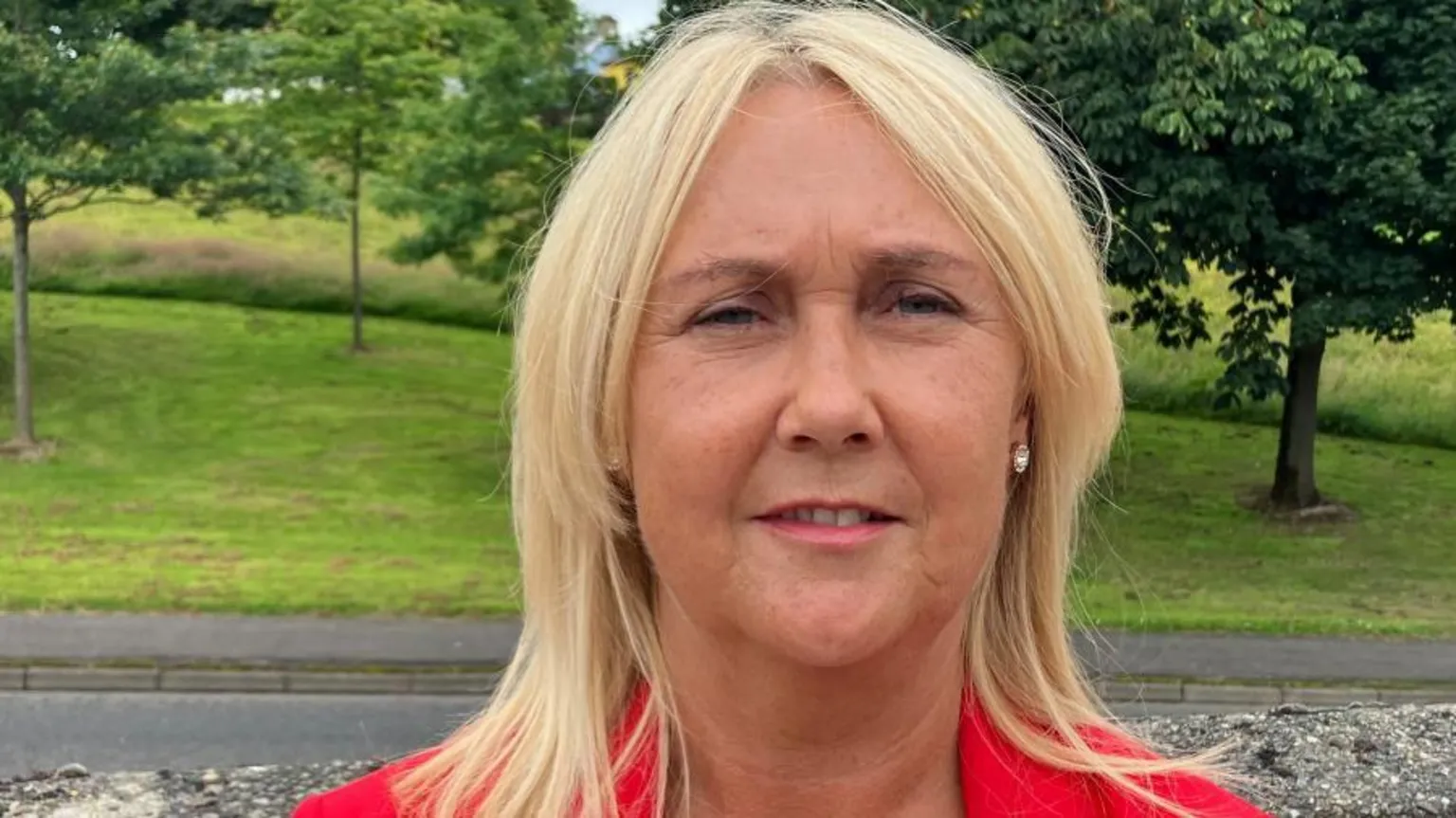 A woman with shoulder length blonde hair looks into the camera. She is wearing diamond earrings and a red jacket with a white top. She is standing in front of a low wall and in the background is a green area with trees.