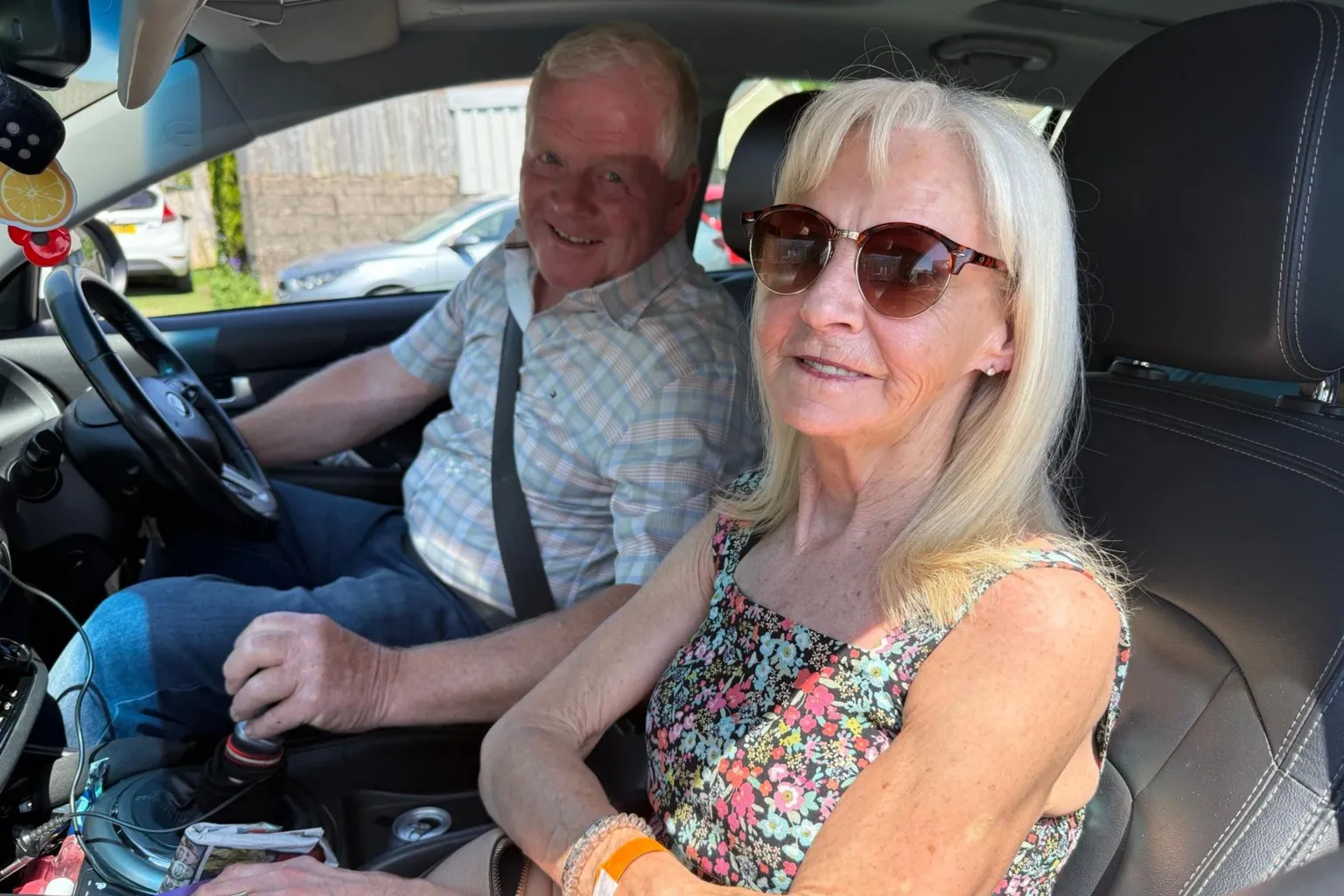 A slim, middle-aged woman with long blonde hair and sunglasses in the passenger seat of a car, next to a man in the driver's seat of a similar age. She is wearing a sleeveless sundress, he wears a checked shirt and jeans. They are both smiling at the camera