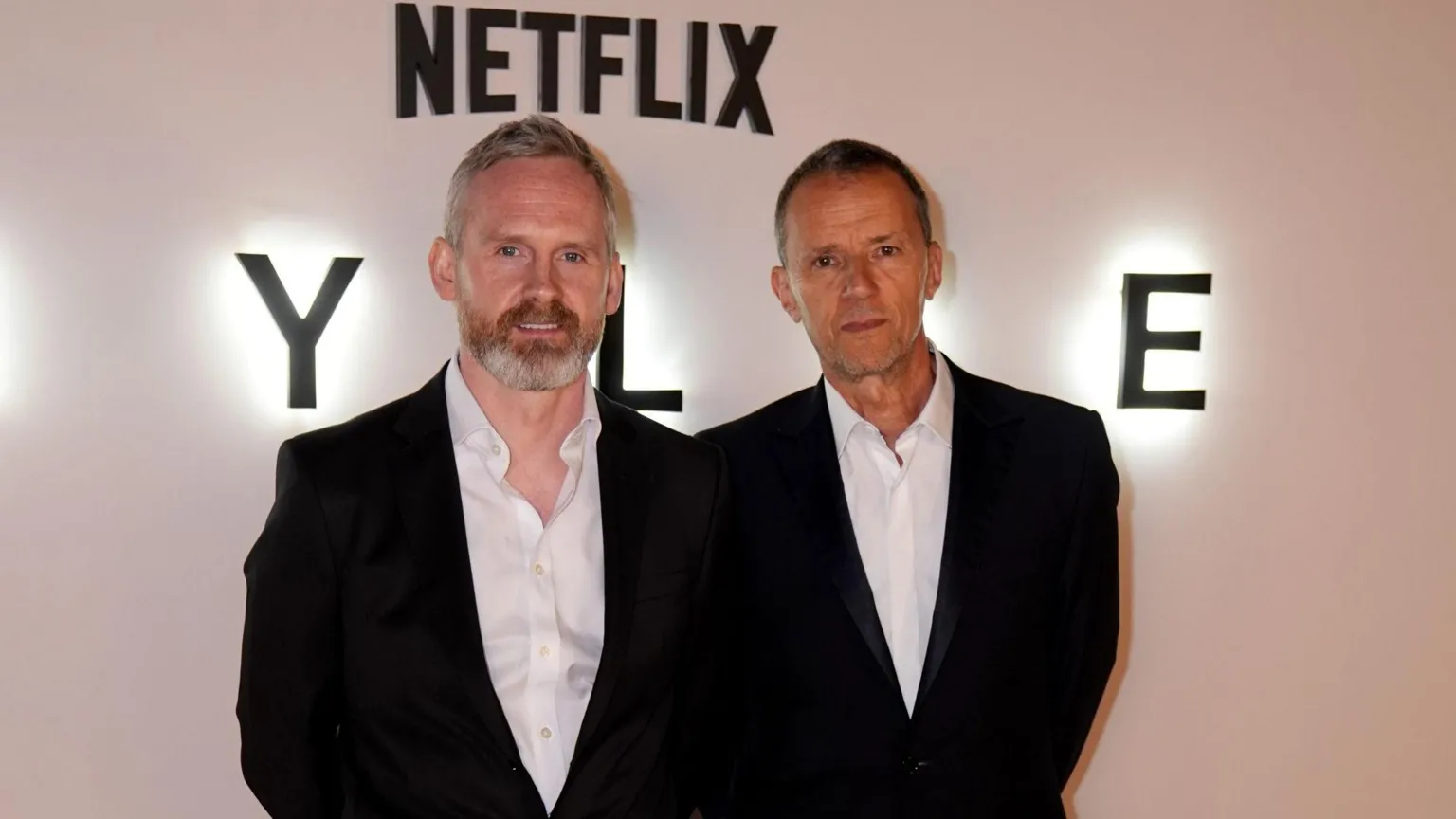  Two men in dark suits and white shirts standing in front of a white wall. Netflix is written on the wall above them.