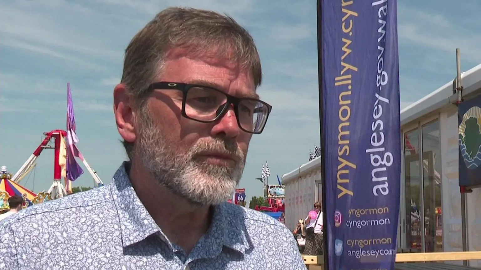 Gary Pritchard doing an interview at the Eisteddfod. He wears a blue patterned shirt in front of Anglesey Council purple branding.