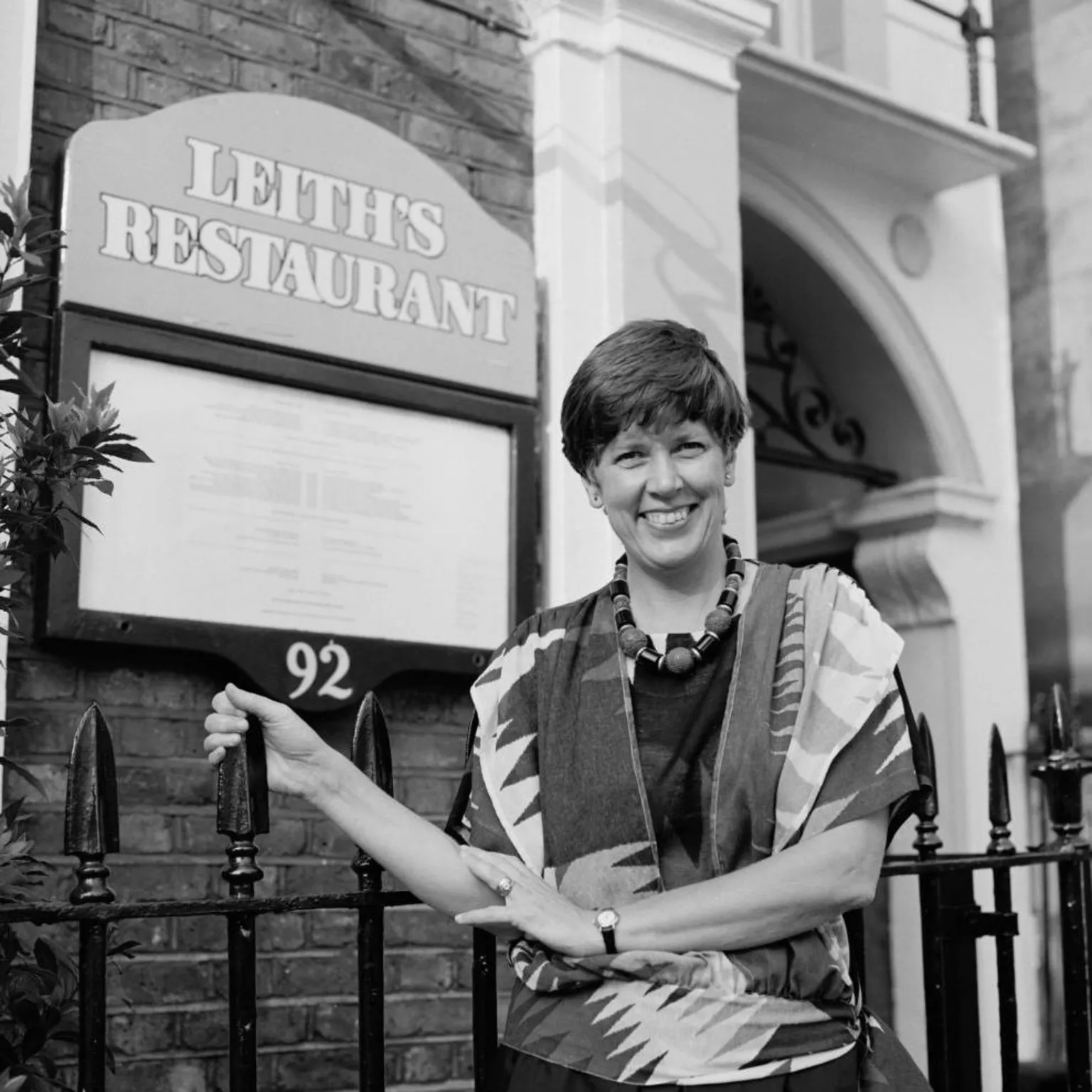  A black and white photo of a woman with short dark hair, wearing a patterned top and chunky beaded necklace, stood in front of a black gate. Behind her is a building with the a board attached which reads 
