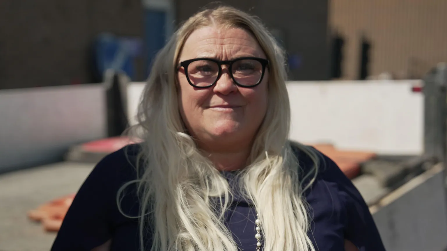 A close-up outdoor portrait of a woman facing the camera. She has long, light-coloured hair and is wearing dark-framed glasses, a dark top, and a long beaded necklace.