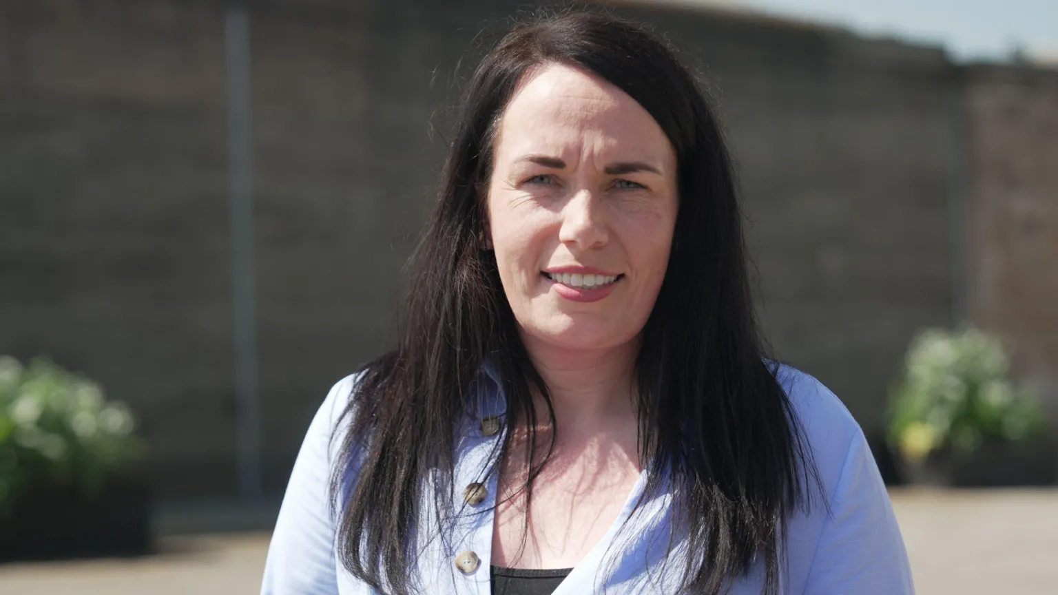 A close-up outdoor portrait of a woman facing the camera. She has long dark hair and is wearing a light blue buttoned top over a darker underlayer.