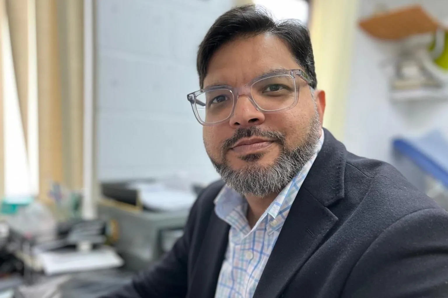 Dr Amir is sat at his desk in his consultation room, looking at the camera with a neutral expression. He has dark hair and a beard and is wearing a checked shirt and dark blazer, with light rimmed glasses. Behind him, out of focus, are office supplies on the desk.
