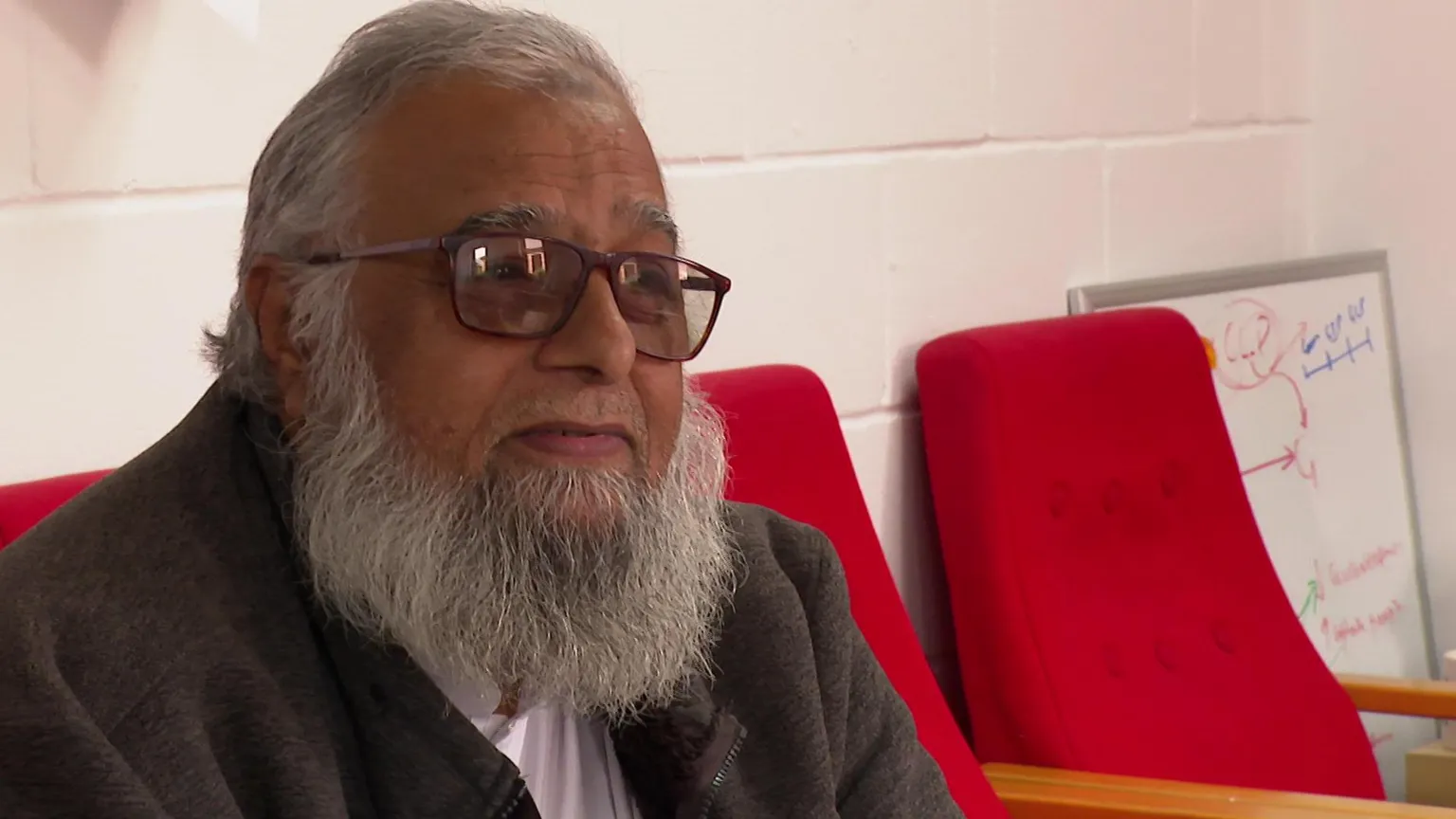 Mahmood is sat on a row of red chairs, he has a grey beard and is wearing dark framed glasses and a dark jacket.