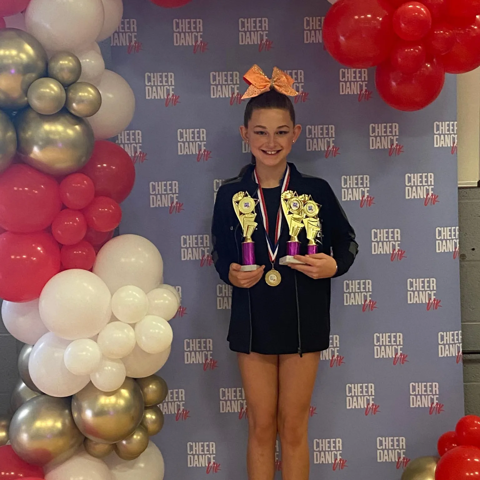 Amanda Axiak A young girl wearing an oversized black sweatshirt. She has her hair tied up with a large orange bow at the front. She has a medal around her neck and is holding three troophies. She is stood in front of a pink and blue background with the words