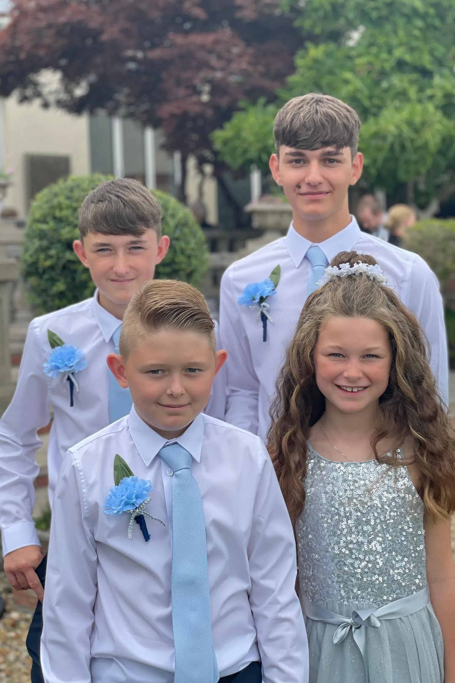 Amanda Axiac Three young boys and a young girl, smiling and looking at the camera. The boys are all wearing white shirts, pale blue ties and a pale blue flower pinned to their chest. The girl is wearing a pale blue sparkly dress and has long wavy brown hair which is partly pulled into a bun. Behind them are trees.