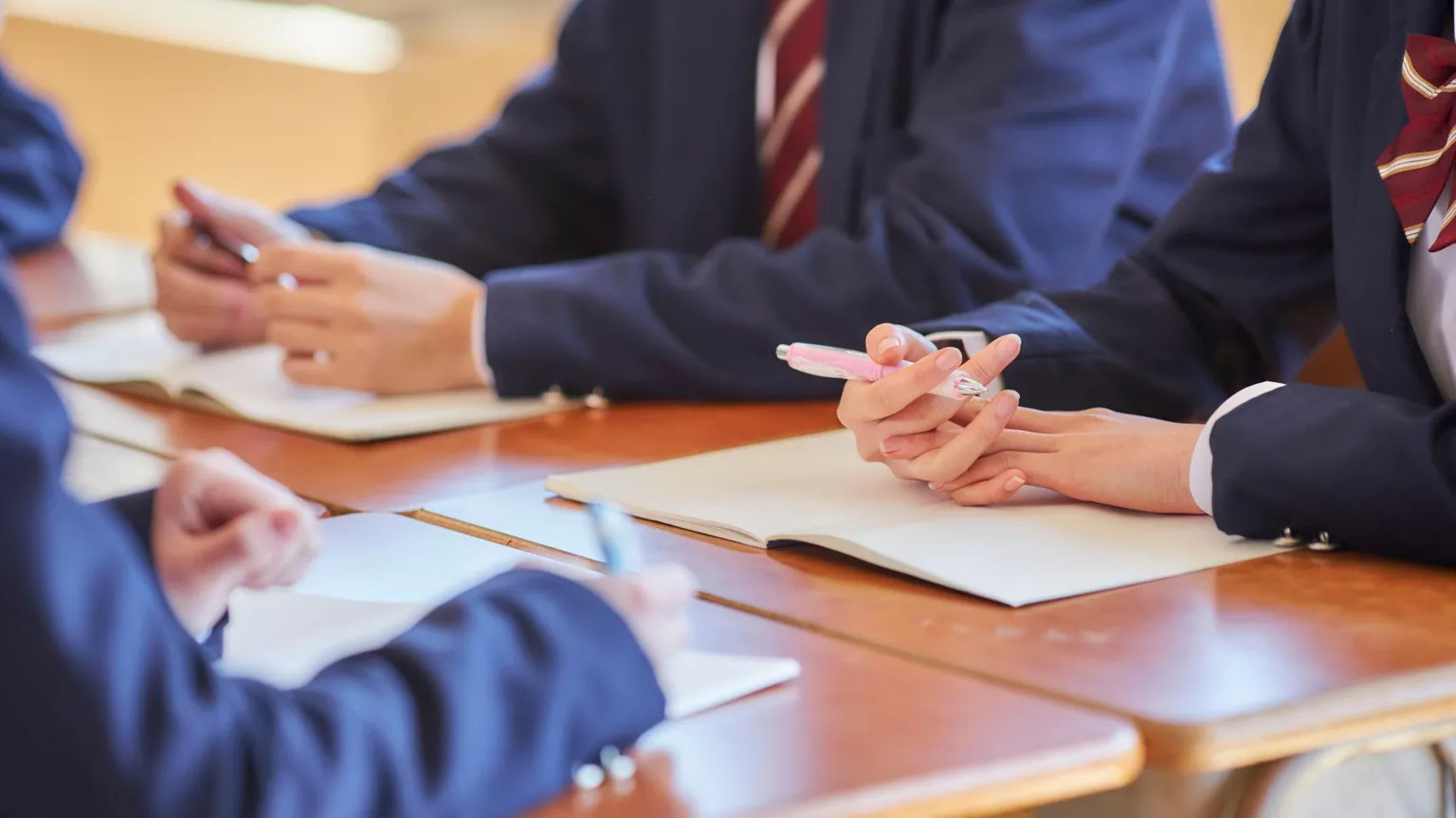  Anonymous students wearing uniforms and sitting at desks. The photo is cropped so only the students' arms are visible. They are wearing navy blazers and burgundy ties.