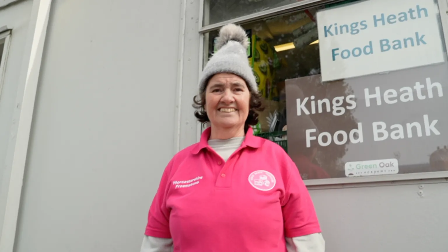 Kings Heath Food Bank coordinator Sharon Power stands outside the storage unit for the charity wearing a pink polo shirt with the food bank's insignia and a sponsor's logo for Worcestershire Freemasons on top of a charcoal grey jumper. On top of her short brown hair, she is wearing a tall, grey bobble hat