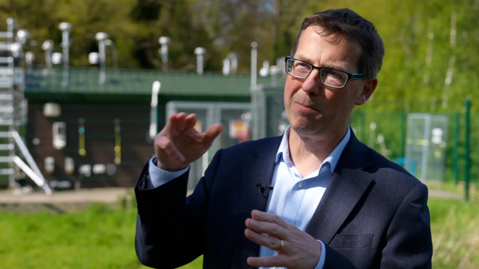 Prof William Bloss wears a navy suit and open collar pastel blue shirt and dark rimmed glasses. He is pictured gesticulating with his hands as he talks outside the university's monitoring station on campus which takes readings of nitrogen dioxide and particulate matter 