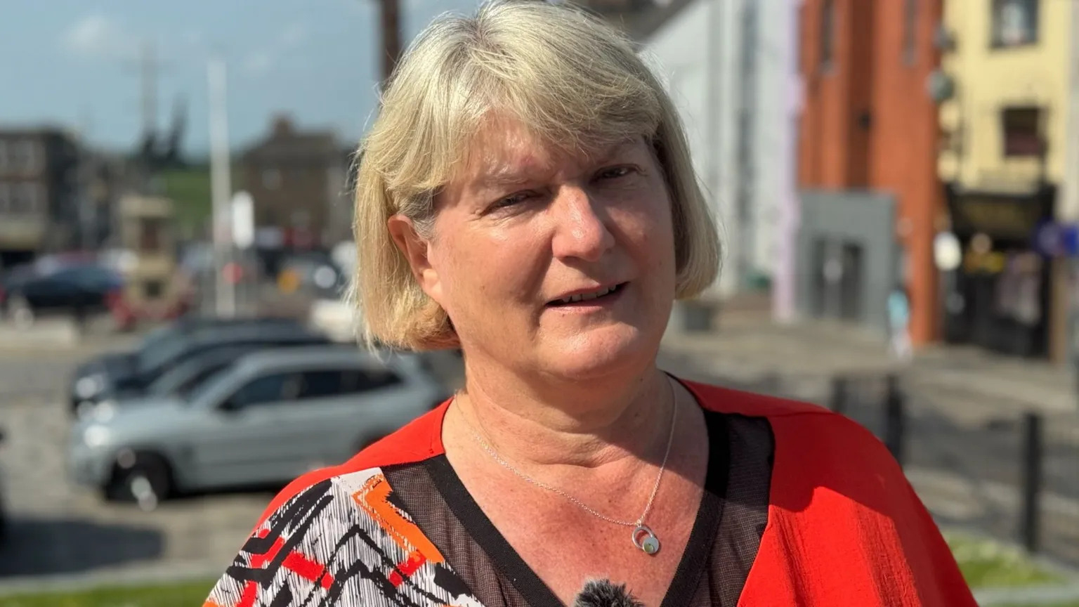 A woman with blonde, bobbed hair looks into the camera, She is wearing a red V-Neck top and has a silver chain around her neck. In the background are parked cars