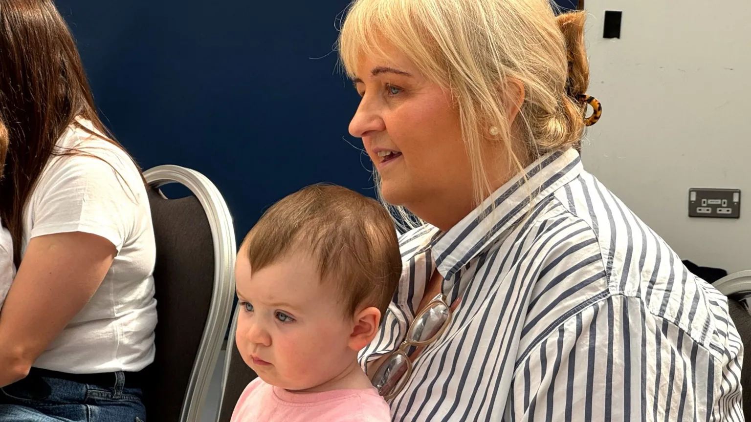 A woman with blonde hair in a bun wearing a white top with black stripes sits on a chair, holding a young child The child has short, dark hair and is wearing a pink top. The side of someone's arm is visible on the right of the pair.