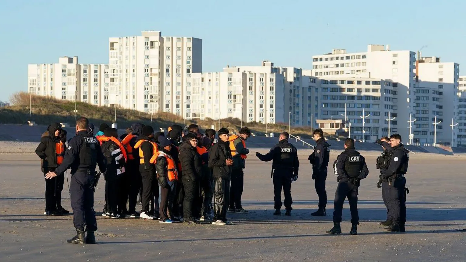  People thought to be migrants are spoken to by police officers after failing to board a small boat off the coast of Dunkirk, France. They are standing on a beach with tower blocks behind them.