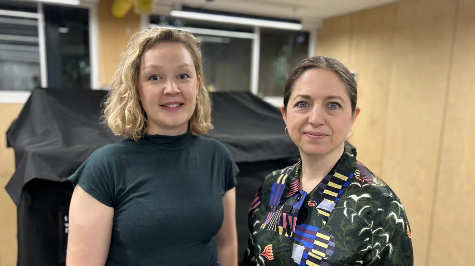 Woman on the left wears a black short sleeved top, has bobbed blonde hair and the woman next to her has a patterned dark blouse and dark brown hair tied back from her face. Behind them are light brown walls.