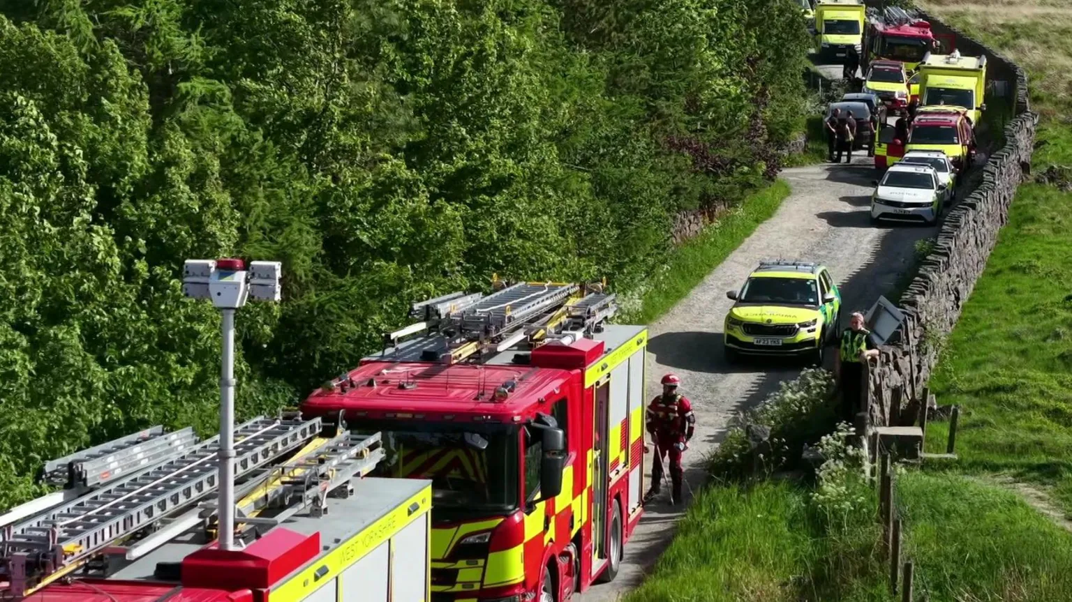 YappApp Emergency services on a rural lane in West Yorkshire. Two fire engines can be seen in the foreground, with a member of the fire crew standing alongside them. An ambulance car can be seen behind these vehicles, along with various parked cars and a number of ambulances.