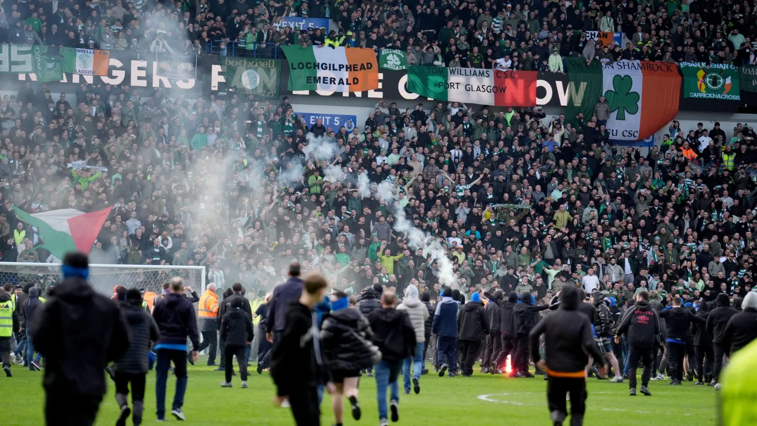 Football fans running across the pitch to confront each other. The majority are clad in black clothing. Celtic fans in the nearest stand, in Celtic tops, are visible watching on.