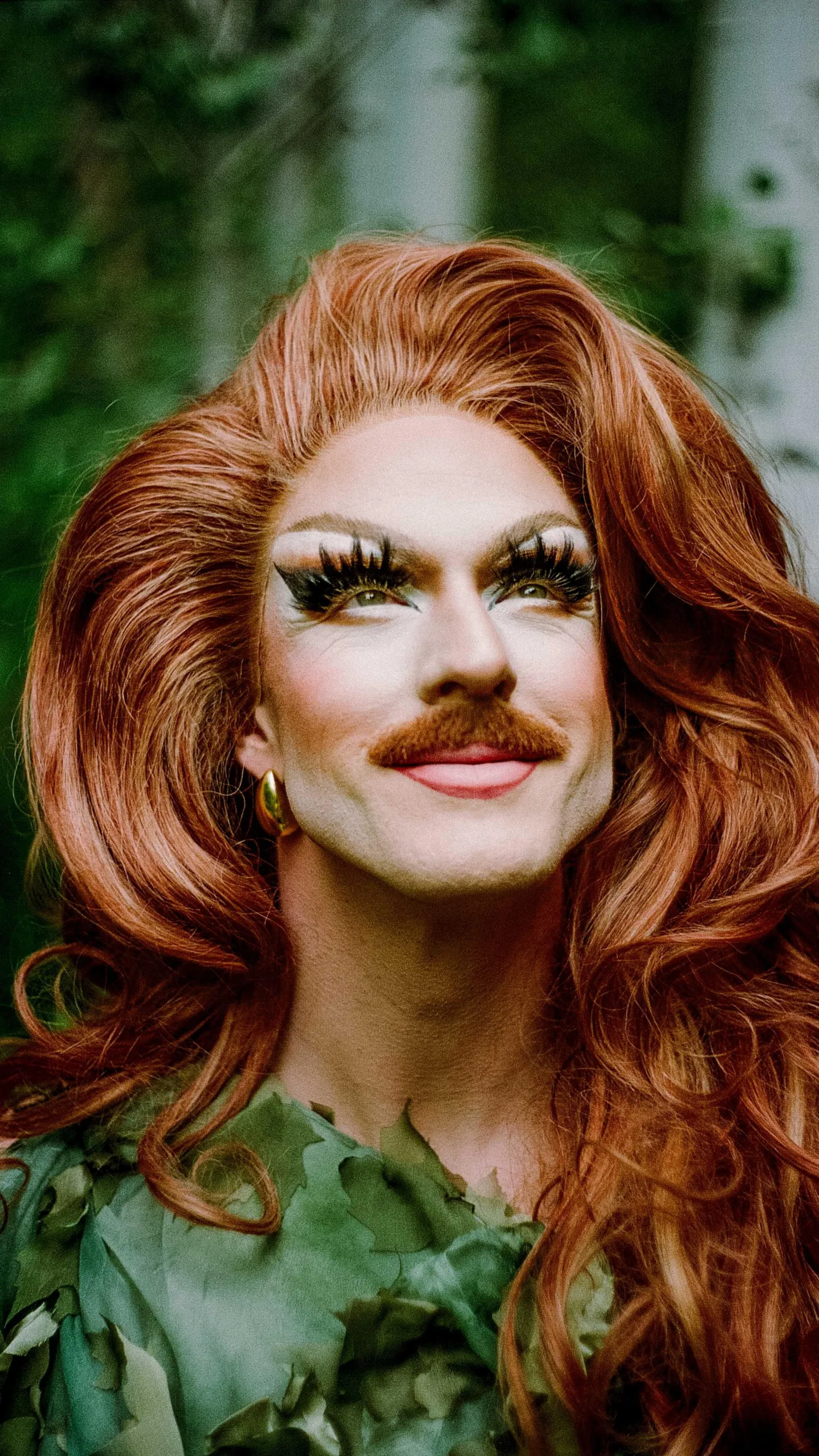 Evan Benally Atwood Pattie Gonia with heavy make up, a mustache, long ginger hair and a green dress on in nature.