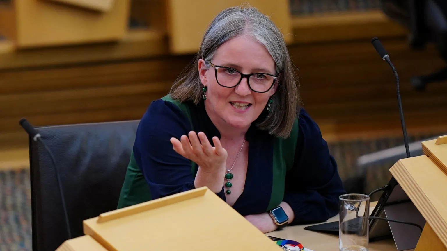  Maree Todd, a woman wearing glasses and with greying hair, sitting at a desk in the Scottish Parliament. She is speaking and has a hand out, gesturing with her palm open.