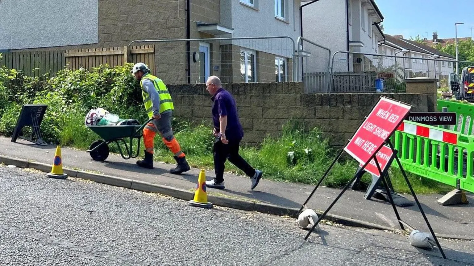 A man in hi vis with a wheelbarrow with household items followed by another man 