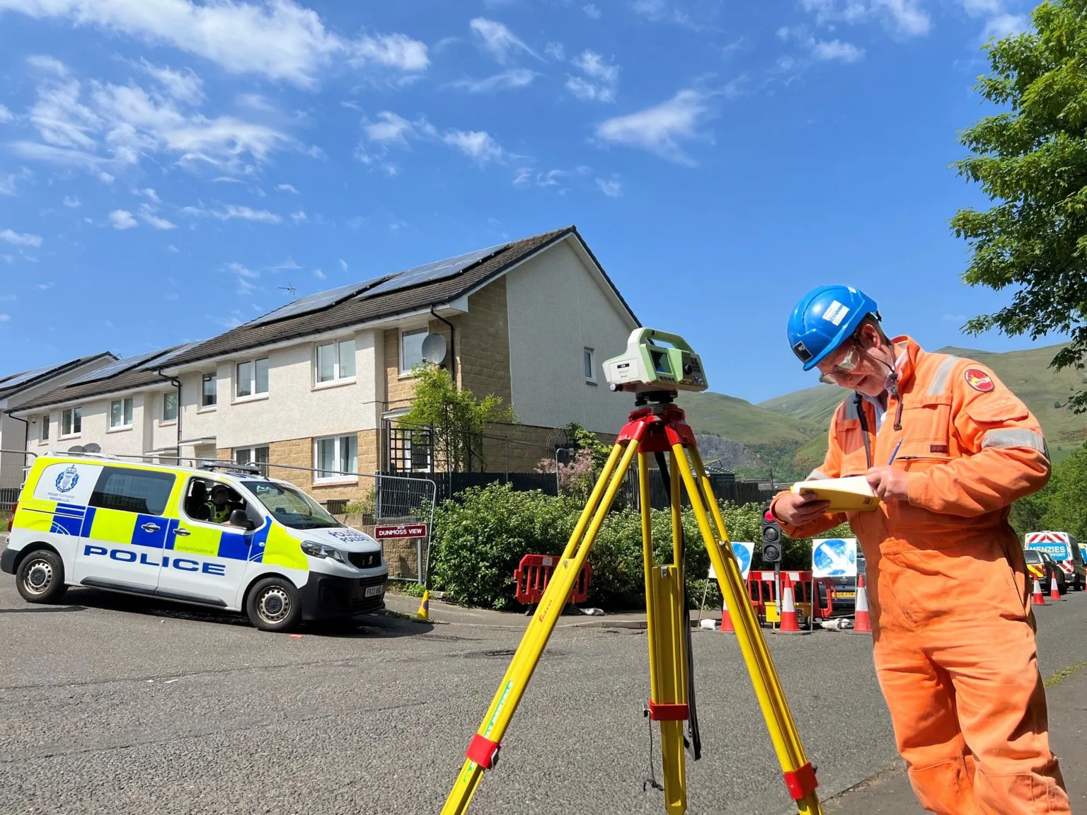A man in hi vis looks at a notebook, with a measuring device beside him. Police officers are visible in the background