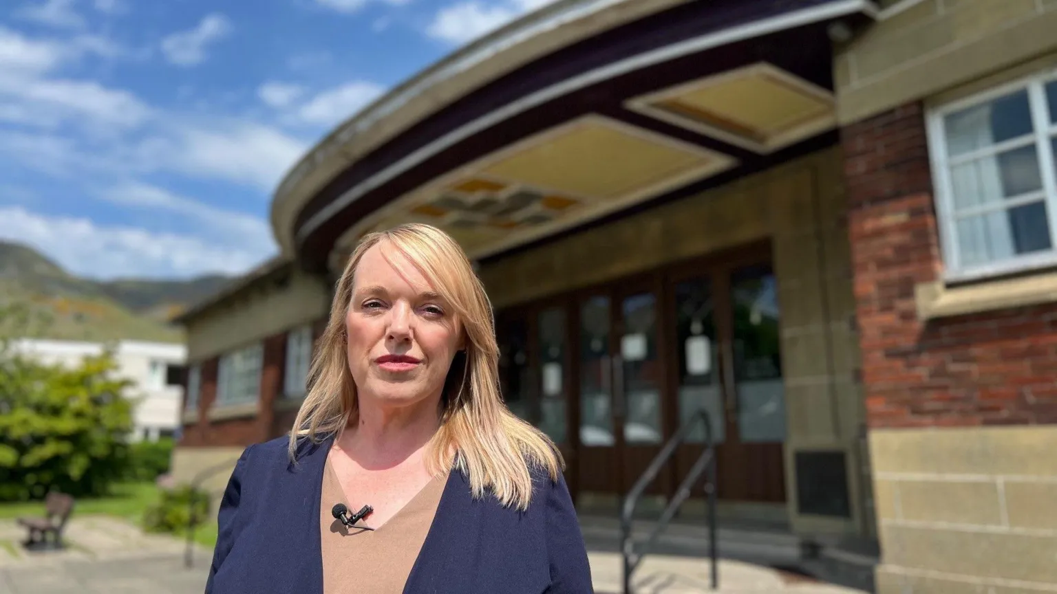 A woman with blonde hair and a blue and brown top stands outside of a community hall