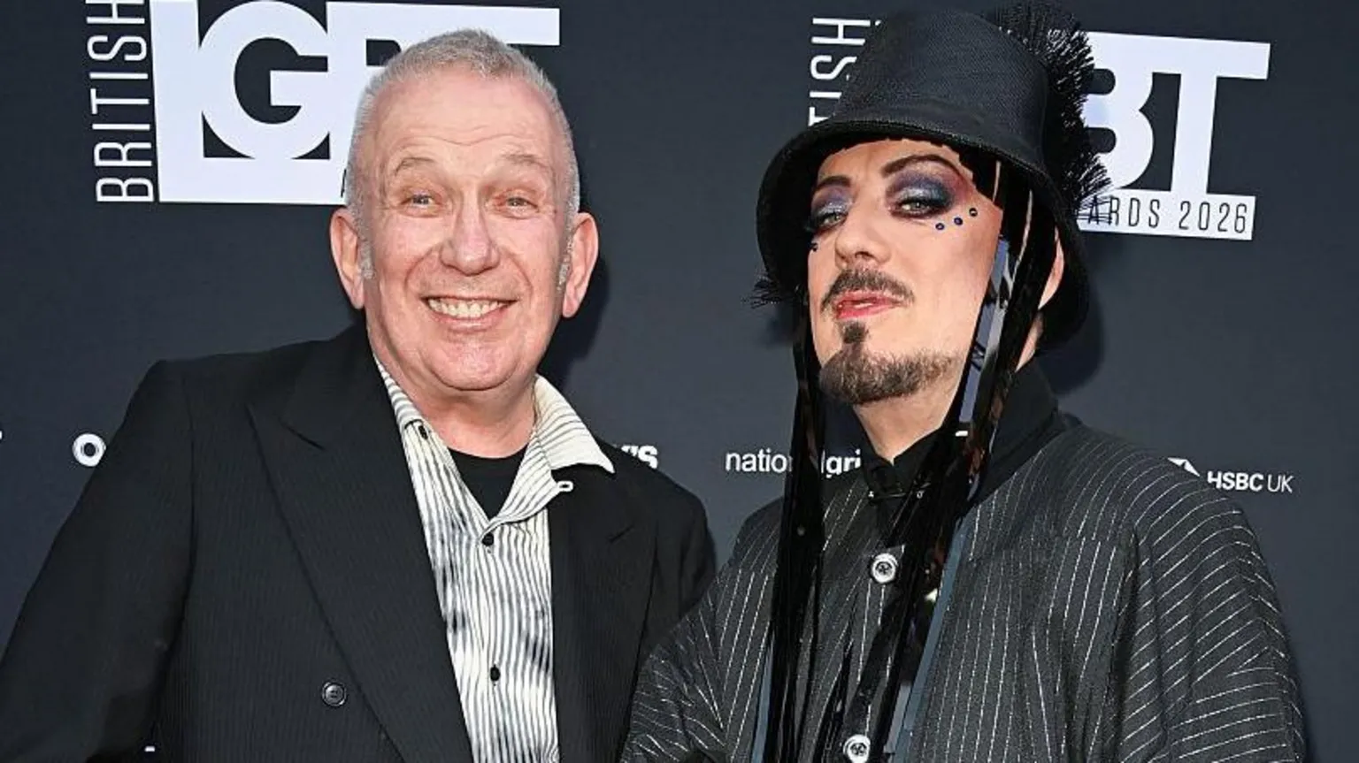 Jean Paul Gaultier and Boy George stand in front of a black wall with the logo 'British LGBT Awards' on it. Jean is wearing a striped white and grey shirt with a black blazer and is smiling. Boy George is wearing a pinstripe jacket and a black bucket-style hat with tassles. He has facial hair and gems around his eyes.