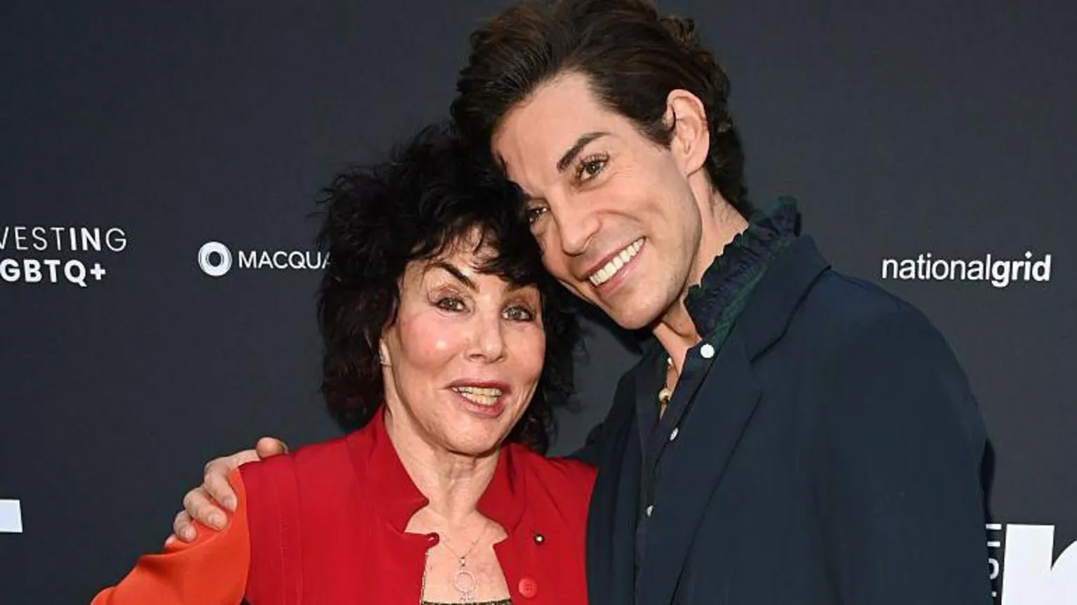 Ruby Wax and Tom Read Wilson pose with their arms around one another. Ruby has black hair and a red jacket with a silver necklace and Tom wears a navy jacket with a blue shirt underneath with a frilled collar. Both are smiling.